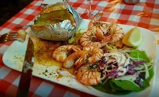 Een bord met gegrilde gamba's, een gepofte aardappel in folie, coleslaw van rode kool en frisse salade op een rood-wit geruit tafelkleed