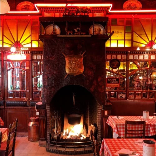 De zaal van Amadeus Gent 1 met een grote open haard, een wapenschild aan de schouw en rood-wit geruite tafels in warm rood licht